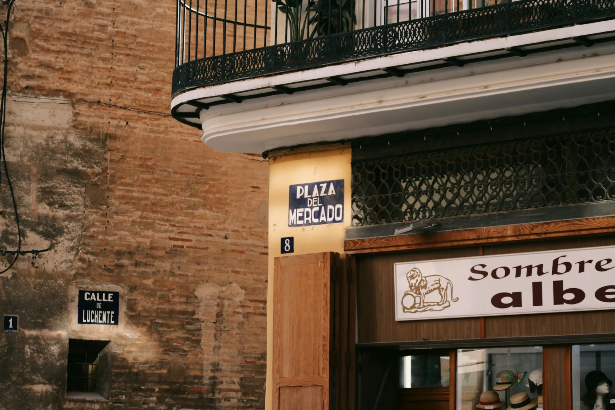 Esquina de una calle española: placas de cerámica con los rótulos «Plaza del Mercado» y «Calle de Luchente» sobre un muro de ladrillo visto, junto al escaparate de una sombrerería.
